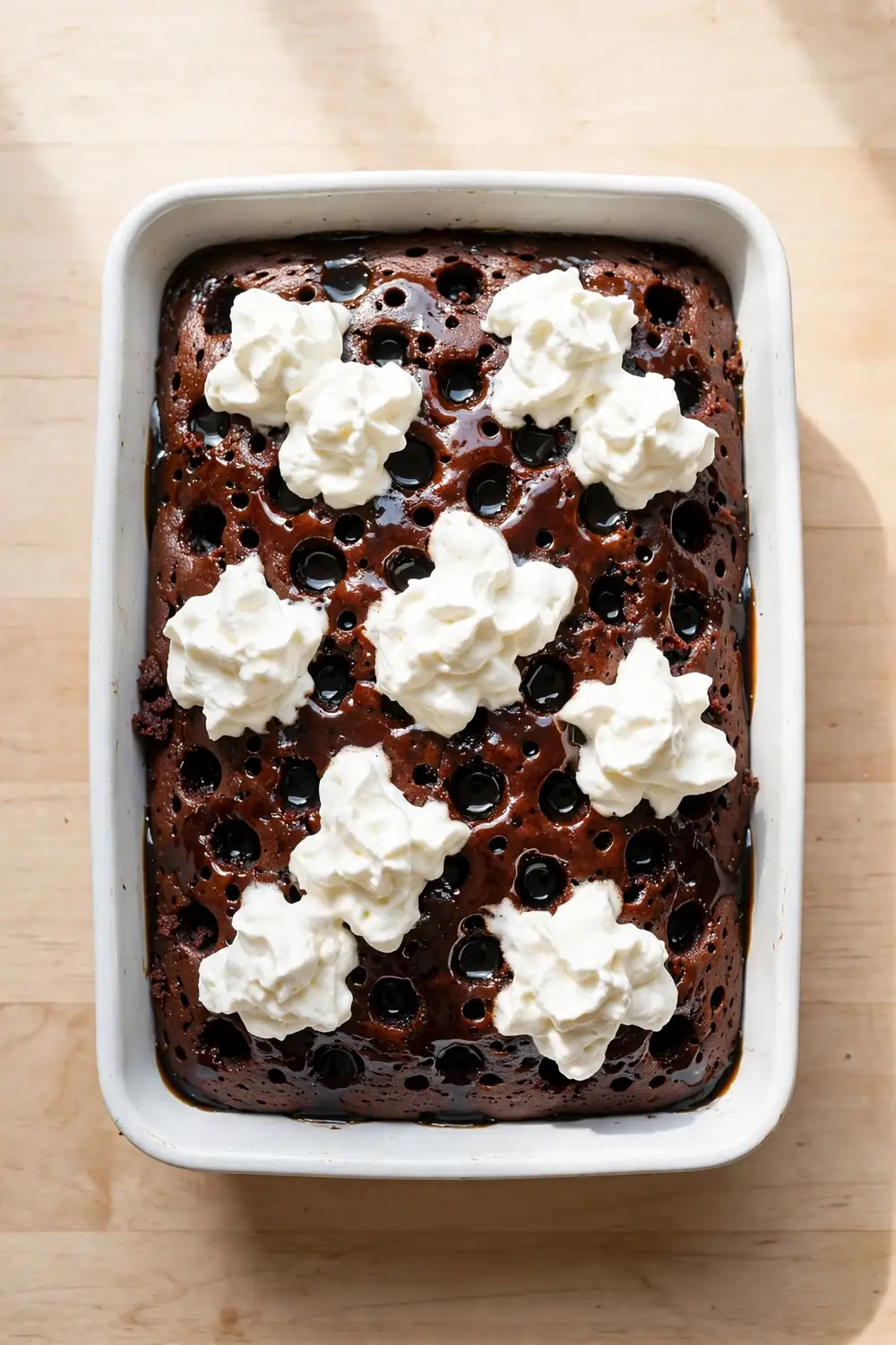 Cola poke cake with glossy cola sauce in poked holes and whipped cream dollops in a baking dish
