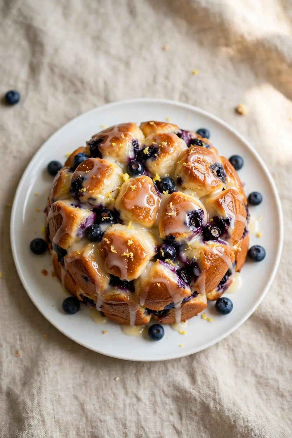 Vertical 2:3 portrait food photograph of lemon blueberry monkey bread on a white plate, golden pull-apart dough balls with burst blueberries and lemon glaze, the plate centered occupying the center 60 percent of the frame with balanced empty space around it, realistic serving and plate proportions, single focal point, warm natural light, photorealistic, no text